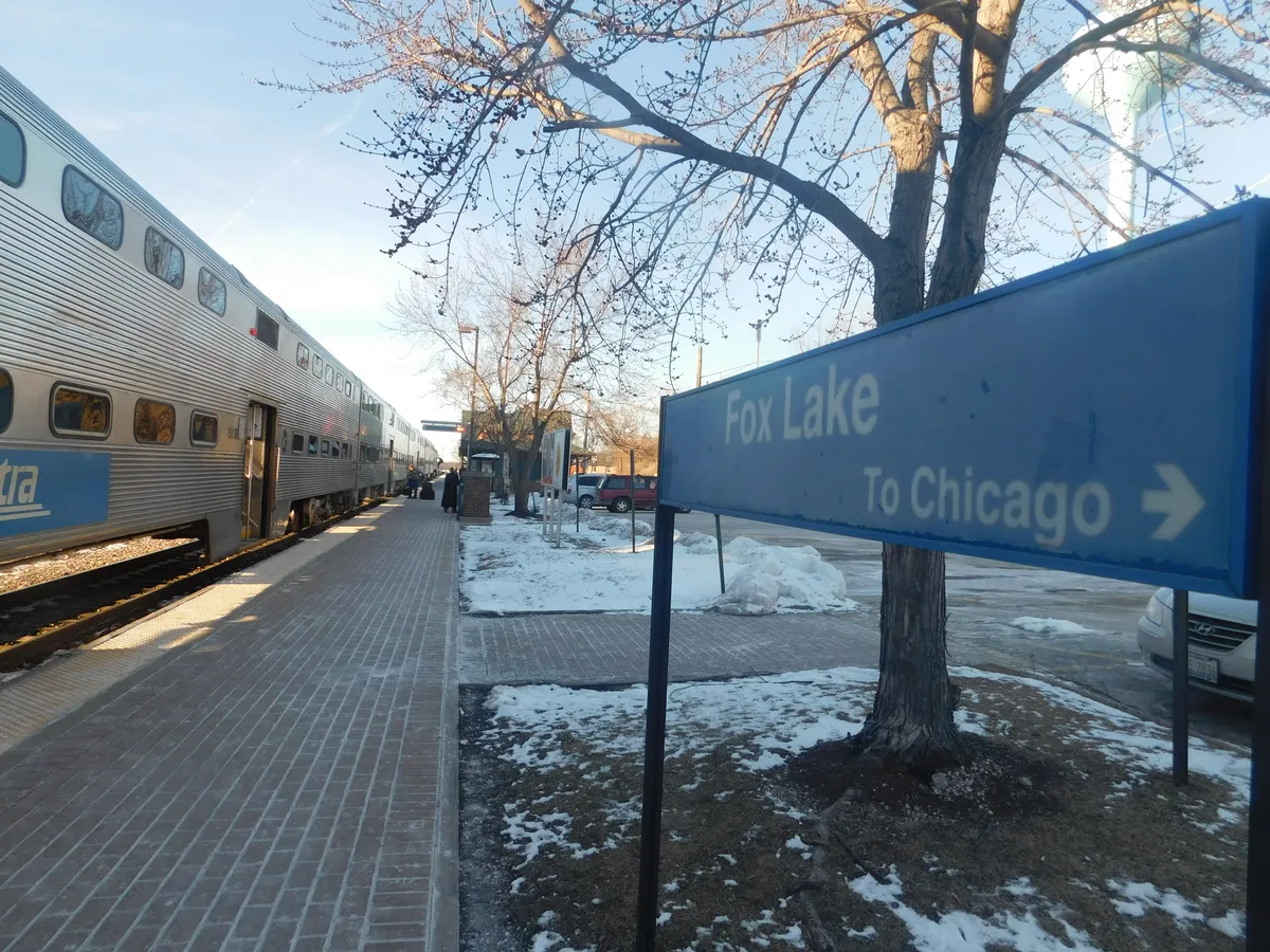 Fox Lake station sign beside a Metra train and a platform lined with bare trees and patches of snow