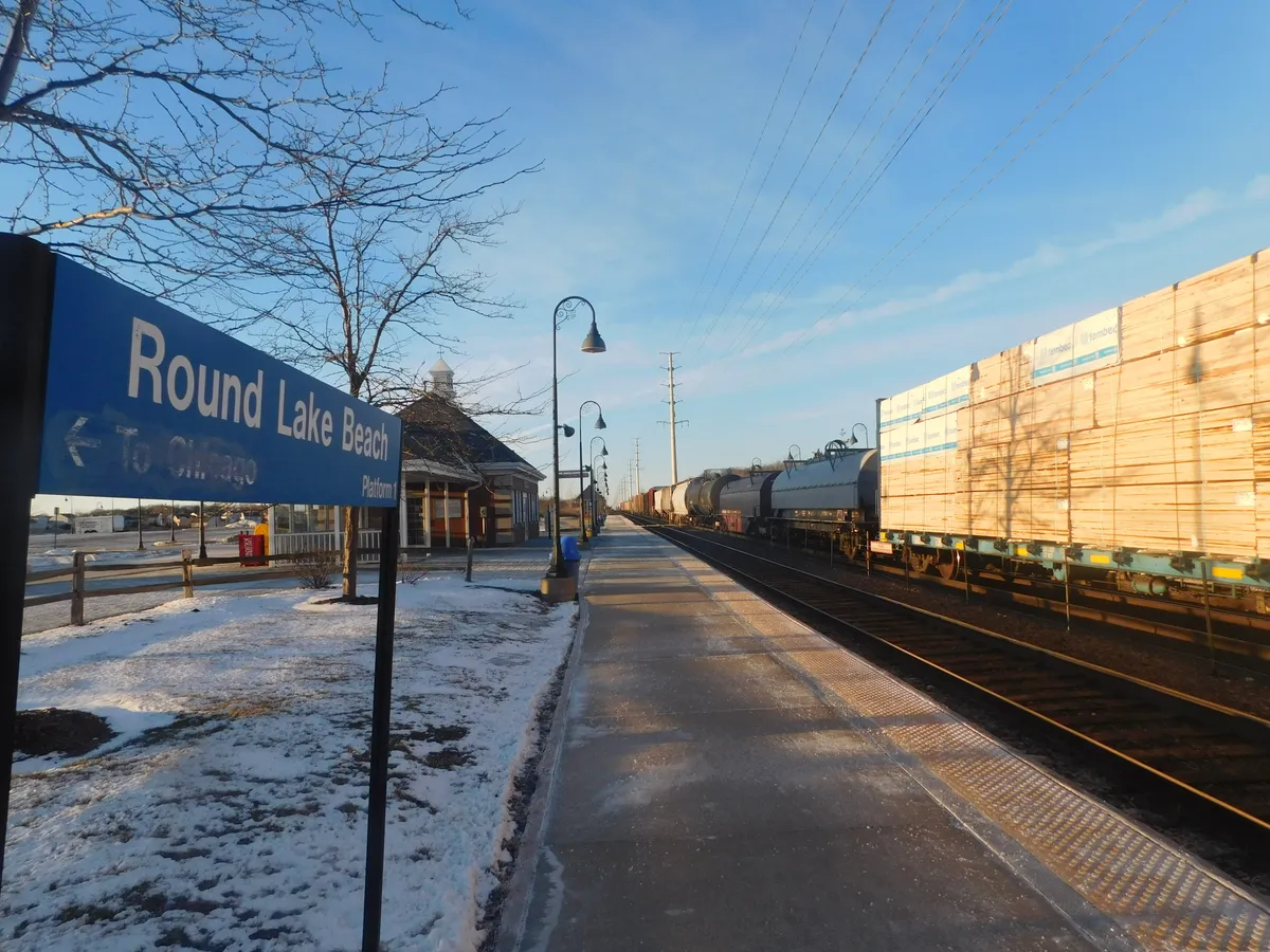 Round Lake Beach station sign and platform beside a freight train, with the station shelter and snow in view